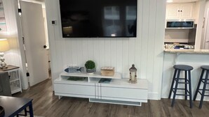 A living room with a flat-screen TV mounted on a white wall, a wooden floor, and a kitchen area with white cabinets and a stainless steel microwave. - Sandpiper Cove 1044 Beach Services  (Destin)