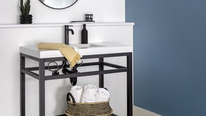 A bathroom with a white sink, black metal frame, a round mirror, a black towel holder, and a woven basket with towels. - Kettle Valley Beach Resort (Penticton)