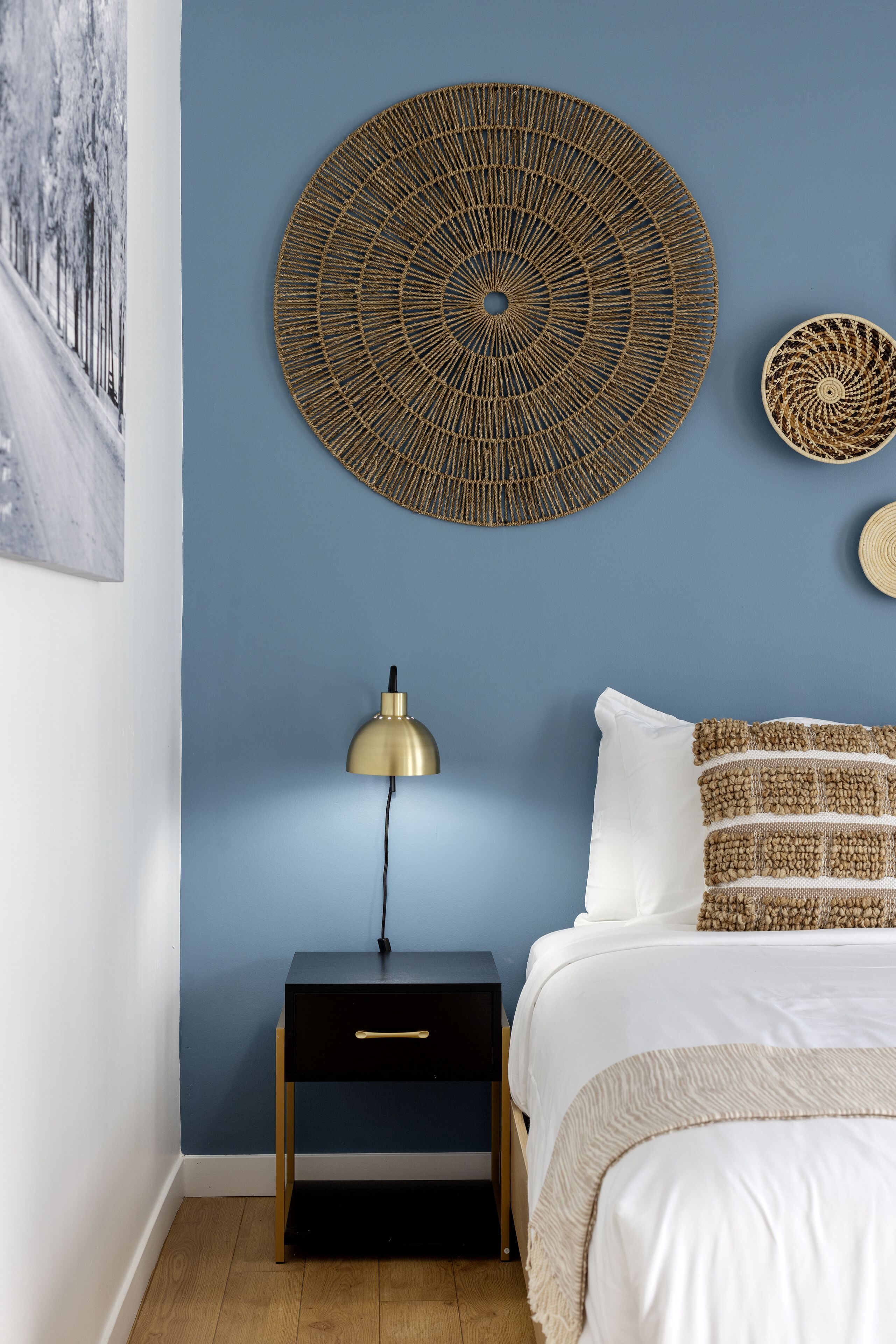 A bedroom with a blue wall, a round woven wall art piece, a bedside lamp, a bed with white bedding and patterned pillows, and a small black nightstand with a drawer.
