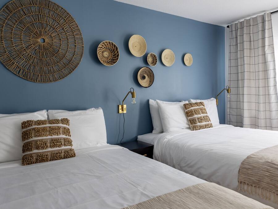 A bedroom with two beds, white bedding, and a blue wall. There are six woven wall hangings and two bedside lamps.