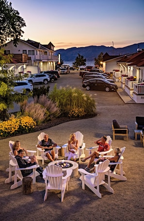 A group of people seated on white Adirondack chairs around a fire pit in an outdoor setting with a view of a lake and mountains in the background. - Kettle Valley Beach Resort (Penticton)