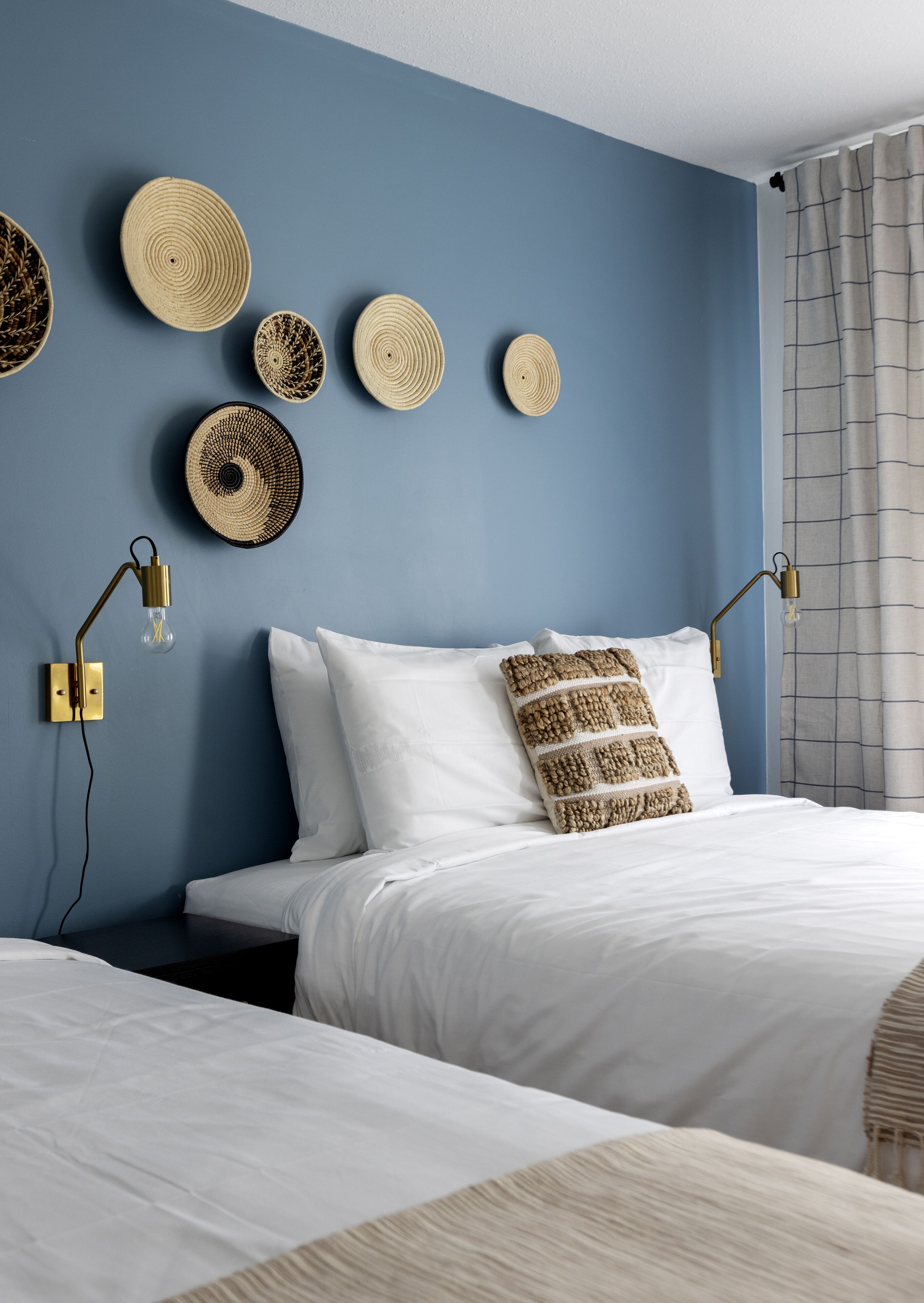 A bedroom with two beds, a blue wall, and woven wall art.
