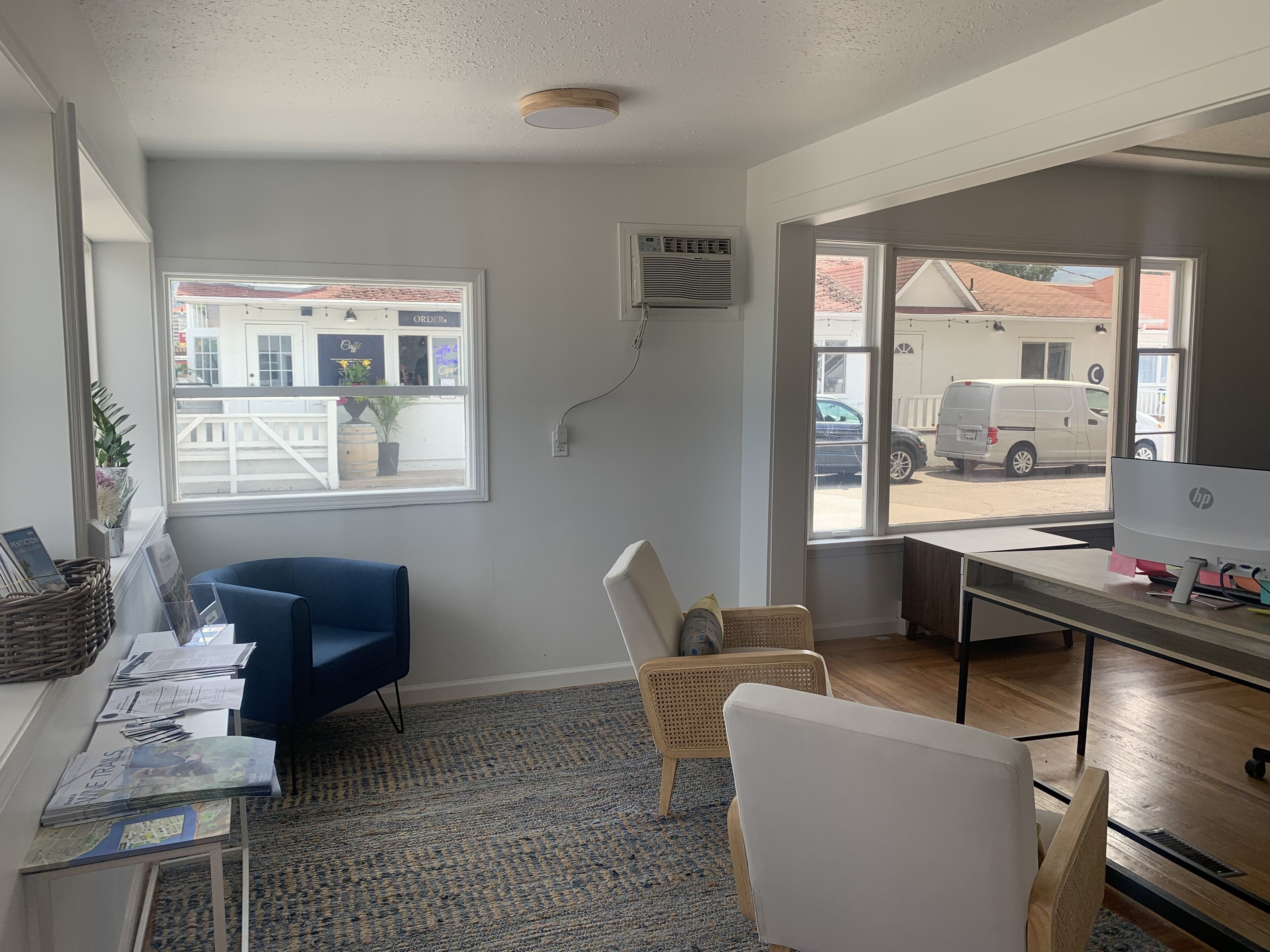 A room with a desk, a blue chair, a wicker chair, a large window with a view of a street, and an air conditioning unit on the wall.