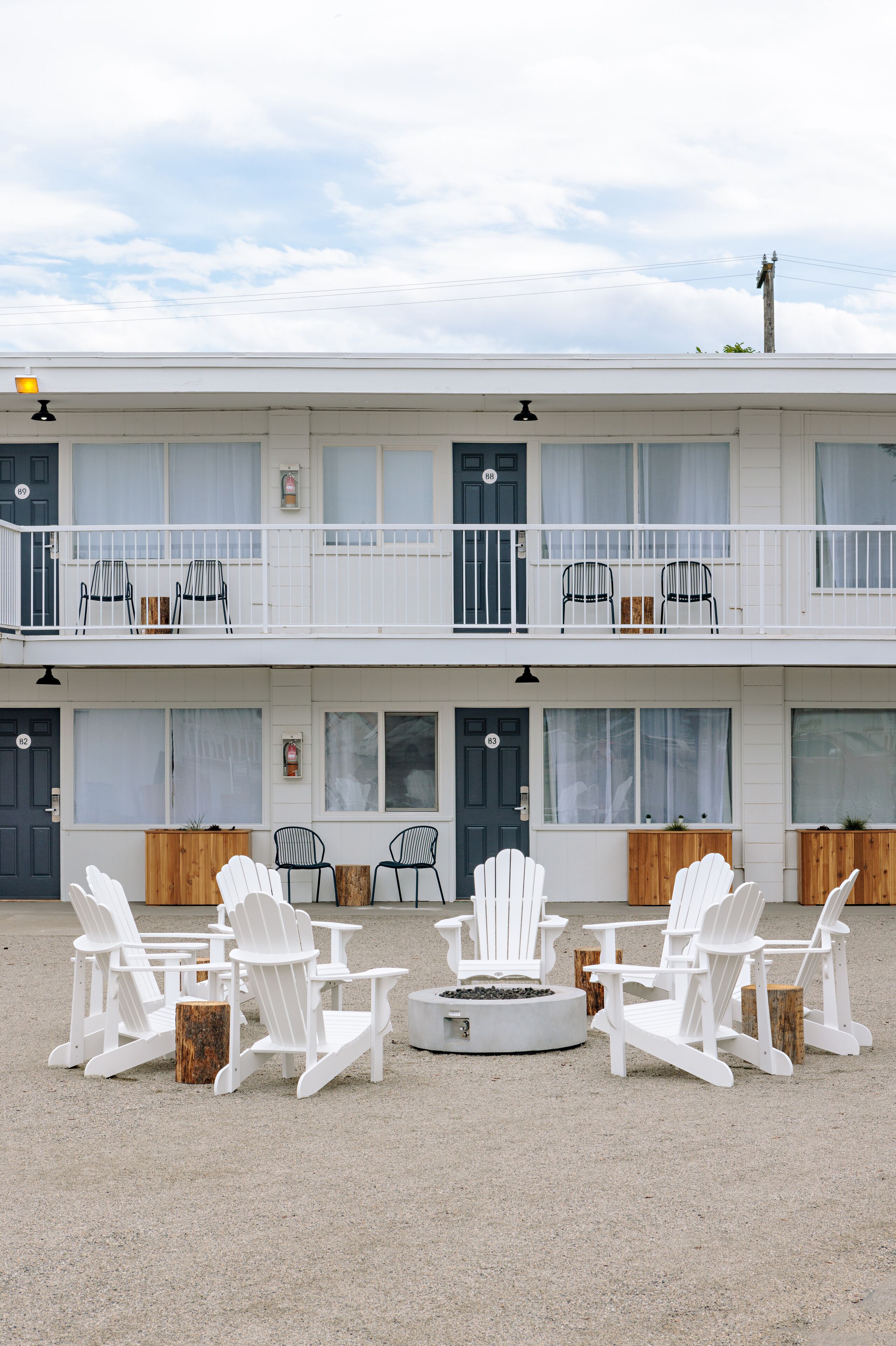 A two-story motel with a balcony on the upper floor, outdoor seating area with white chairs, and a fire pit in the center.