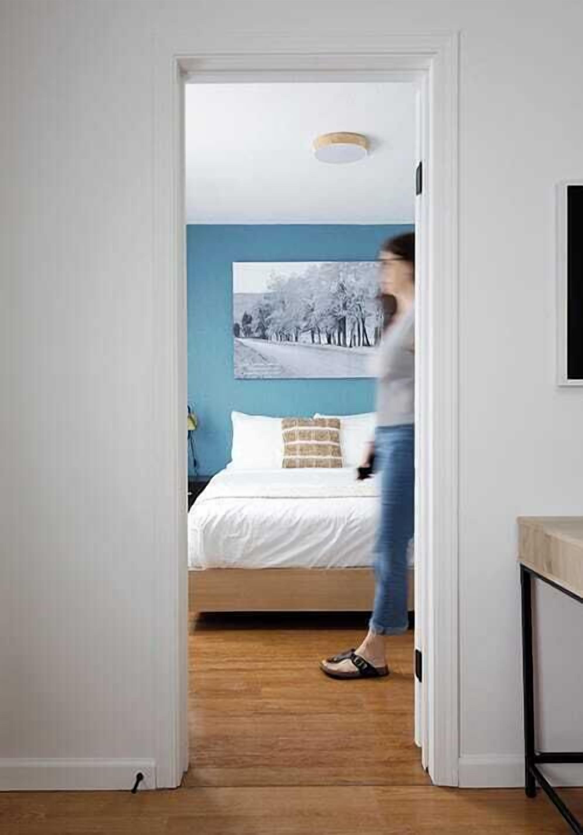 Une porte qui mène à une chambre à coucher, où l’on aperçoit un lit, un tableau accroché au mur et un luminaire au plafond.
