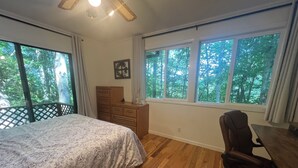 Une chambre à coucher comprenant un lit, une commode, un bureau et une chaise. De grandes fenêtres offrent une vue sur les arbres.