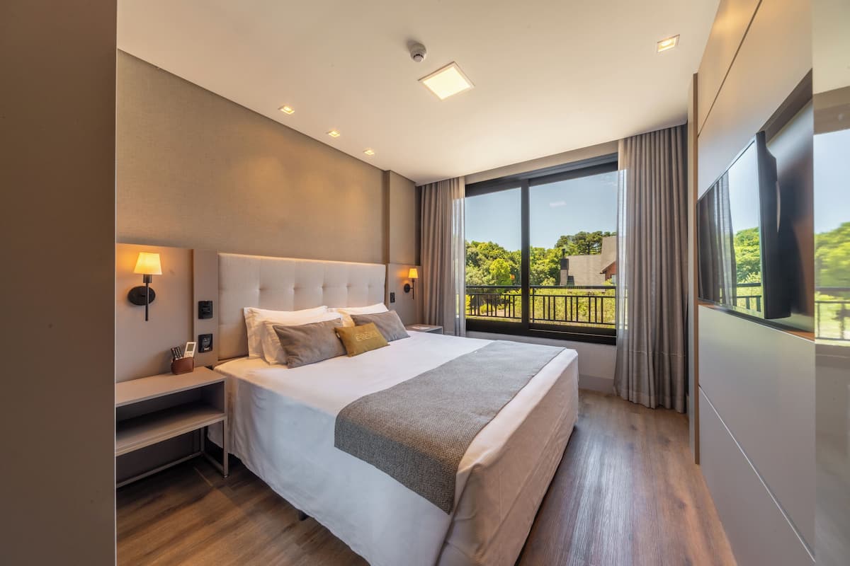 Une chambre d’hôtel moderne dotée d’un grand lit, de tables de chevet, d’une petite table, d’une vue sur la verdure grâce à de grandes fenêtres et d’un balcon.