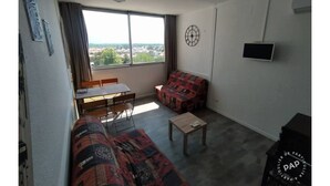 A small, sparsely furnished room with a large window, a sofa, a small table, and a television.