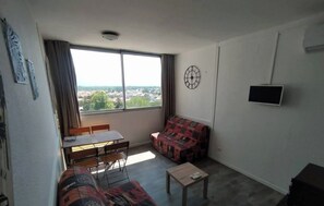 Une petite pièce au mobilier minimaliste, avec une grande fenêtre, un canapé, une petite table et un téléviseur.