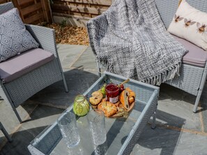 Mesa con cubierta de vidrio y bandeja de comida, que incluye pasteles y una botella de mermelada, ubicada en un patio exterior con mobiliario de mimbre.