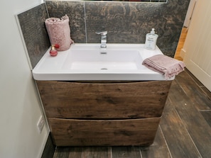 Un baño con un lavamanos blanco, un tocador de madera, una toalla rosa y una botella de jabón para manos.