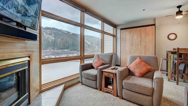 Ein gemütliches Wohnzimmer mit Kamin, zwei Sesseln, einem kleinen Tisch und einem großen Fenster mit herrlichem Blick auf eine verschneite Landschaft.