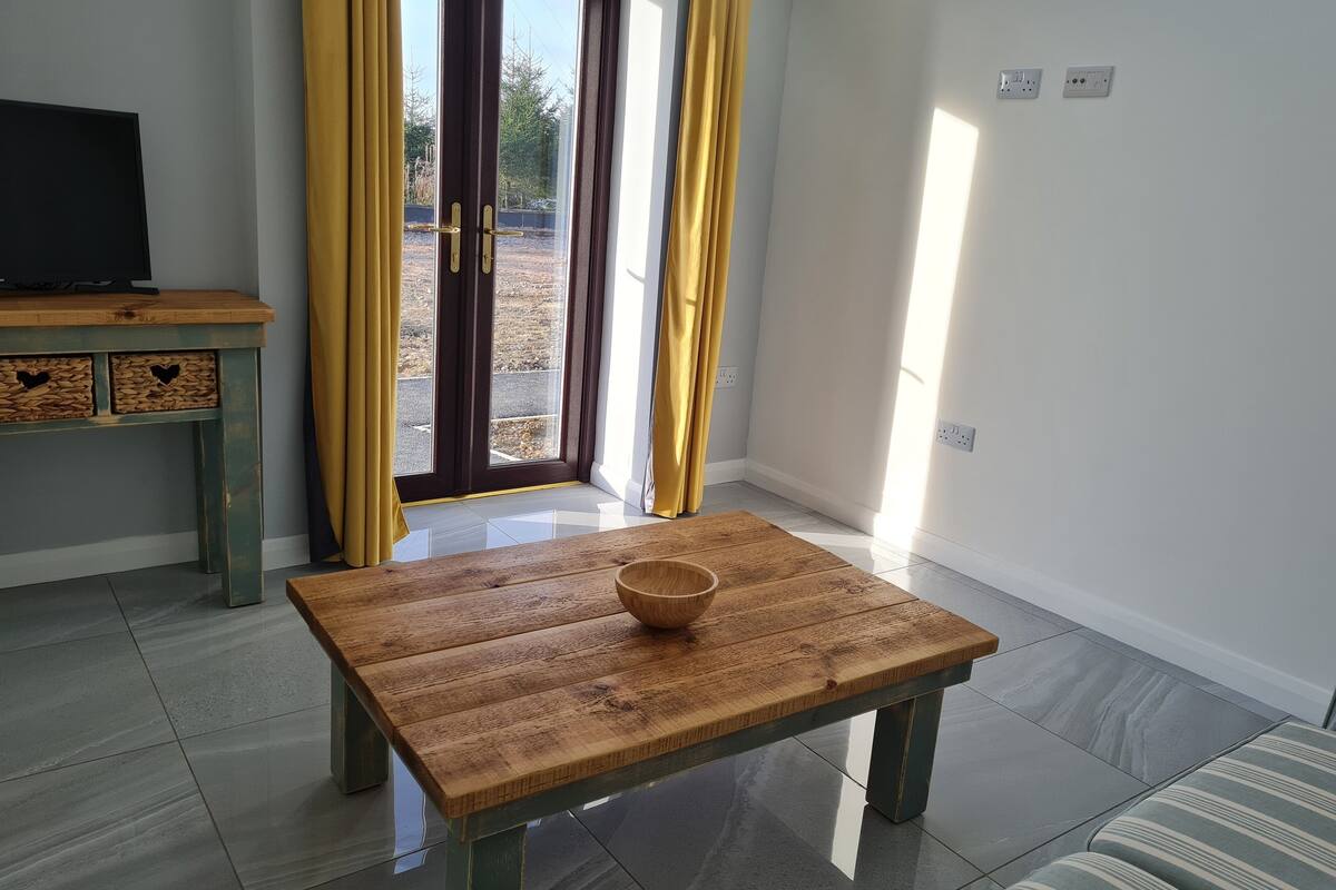 A room with a wooden table, a television on a stand, and a view through a glass door with yellow curtains.