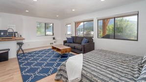 A modern, well-lit room with a large bed, a sofa, a coffee table, and a view of the outdoors through large windows. - Lakeside Bungalows Unit 1&2 (Bayview)