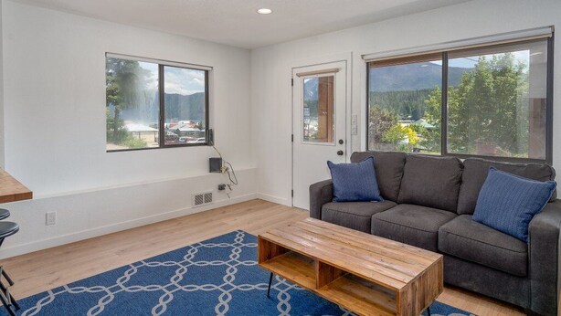 A modern living room with a grey sofa, a wooden coffee table, and a blue patterned rug. There are two large windows offering views of a mountainous landscape and a parking area. - Lakeside Bungalows Unit 1&2 (Bayview)