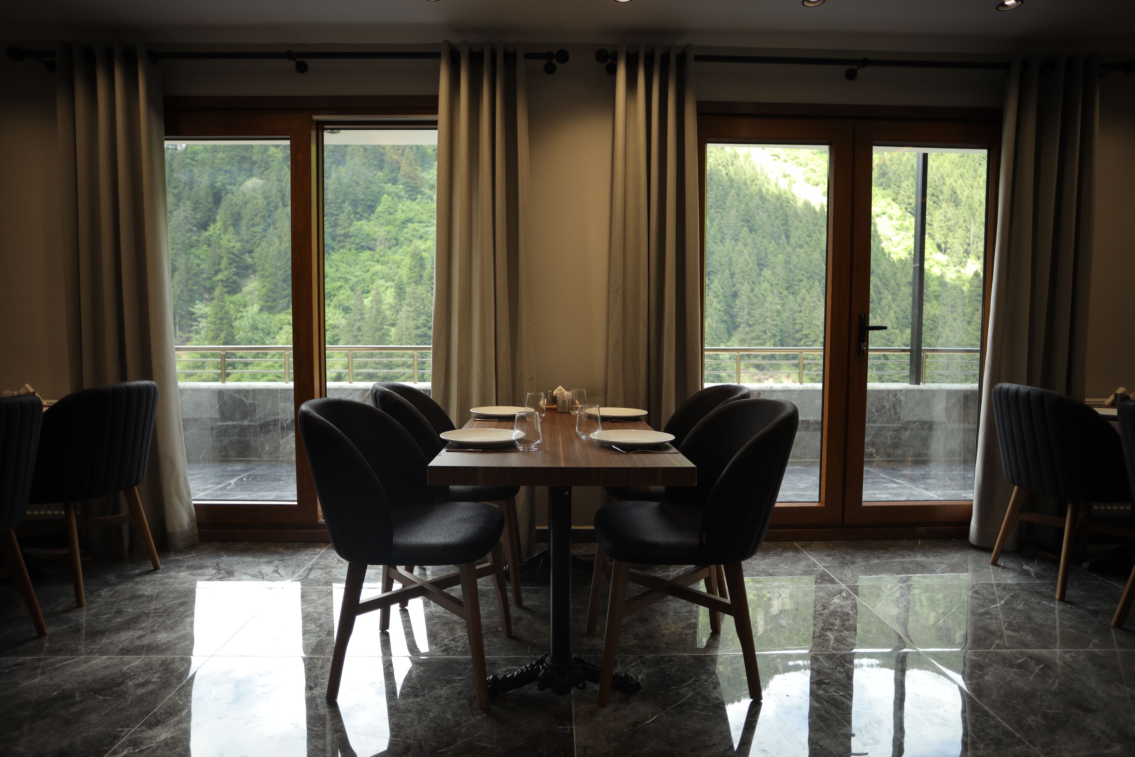 A dining area with a table set for six, surrounded by chairs, large windows with curtains, and a view of a forested landscape.
