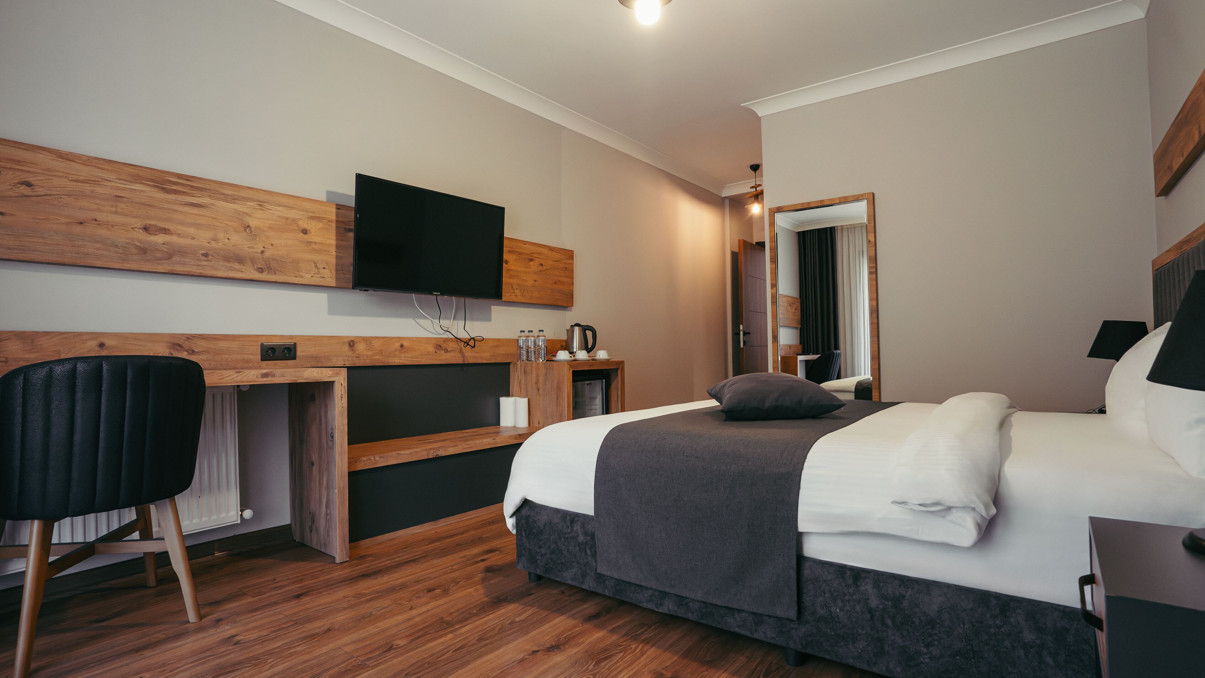 Una habitación de hotel moderna con un escritorio de madera, un televisor de pantalla plana, una cama con edredón gris y un espejo.