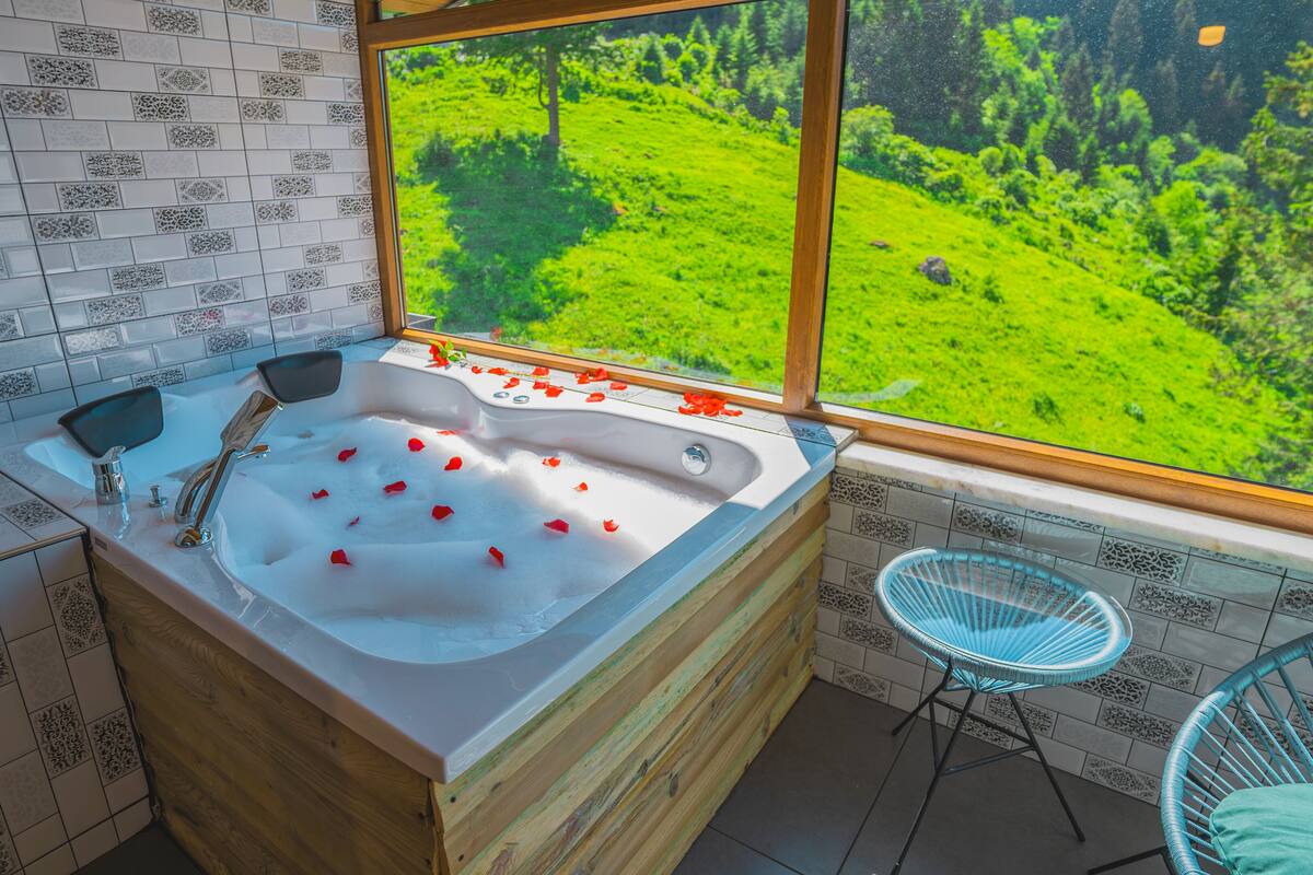 A bathroom with a large bathtub, a wooden cabinet, a blue chair, and a view of a green landscape through a window.