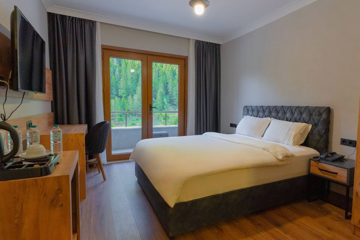 A hotel room with a bed, a desk with a kettle, a chair, a nightstand, and a view of a balcony with curtains.
