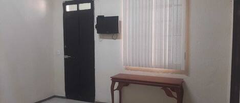 A room with a closed black door, a small wooden table, and a window with blinds.