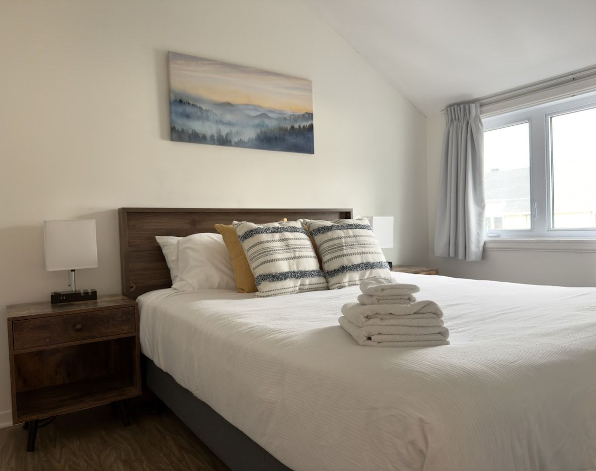 Ein Hotelzimmer mit Bett, Nachttisch, Lampe, einem Gemälde an der Wand und einem Fenster mit Vorhängen.