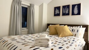 Een net opgemaakt bed met een dekbedovertrek met veermotief, een raam met gordijnen en drie ingelijste kunstwerken met bladeren aan de muur.