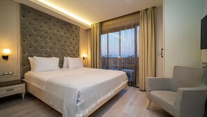 A hotel room with a large bed, a grey upholstered headboard, two bedside lamps, a grey armchair, and a view of the outdoors through sheer curtains. - Rooms365 (Fethiye)