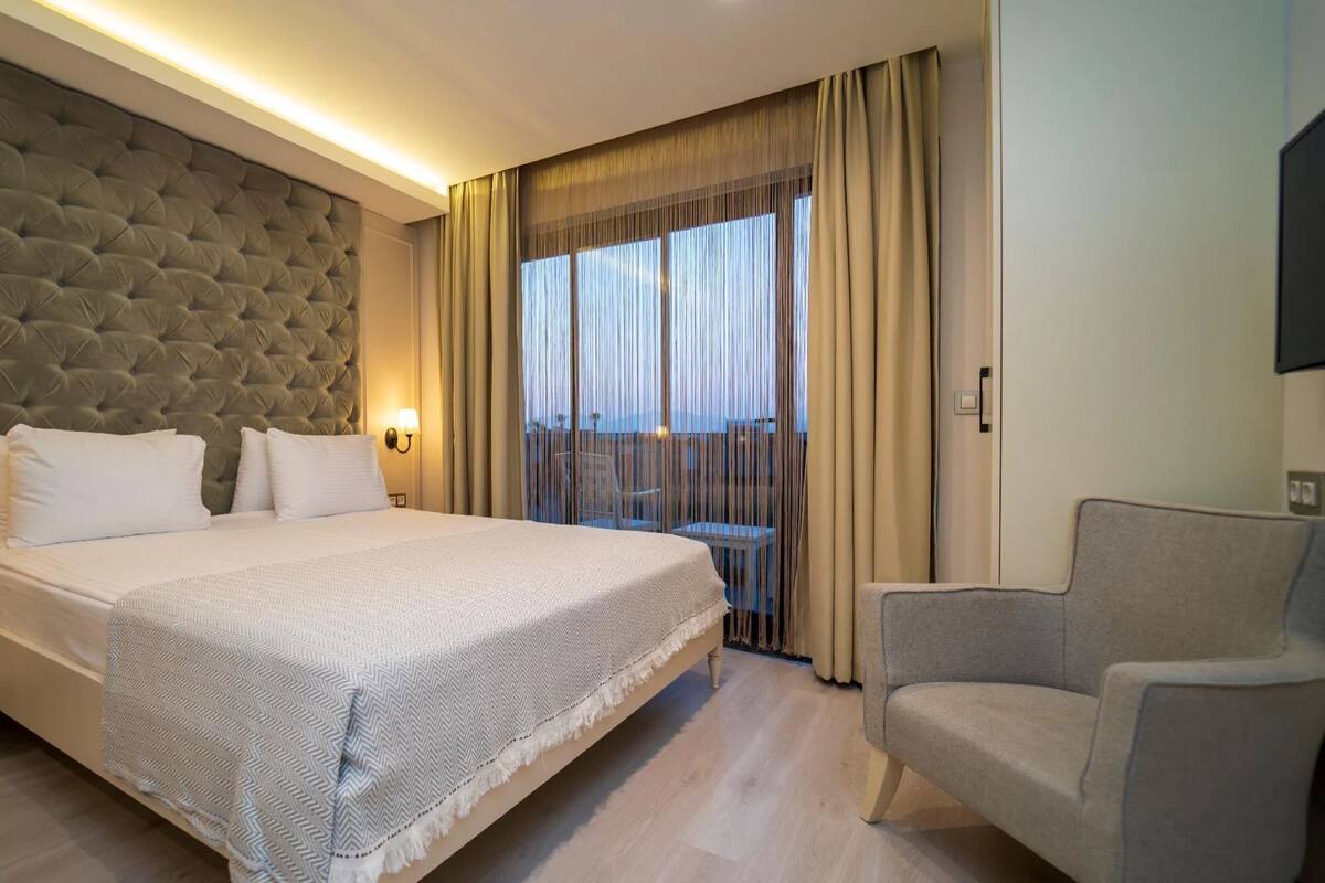A hotel room with a large bed, a grey upholstered headboard, two bedside lamps, a grey armchair, and a view of the outdoors through sheer curtains.