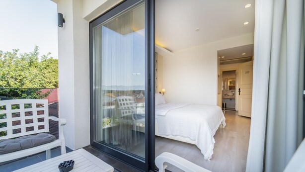 A balcony with a white table and bench, a sliding glass door leading to a bedroom with a bed and a bathroom visible. - Rooms365 (Fethiye)