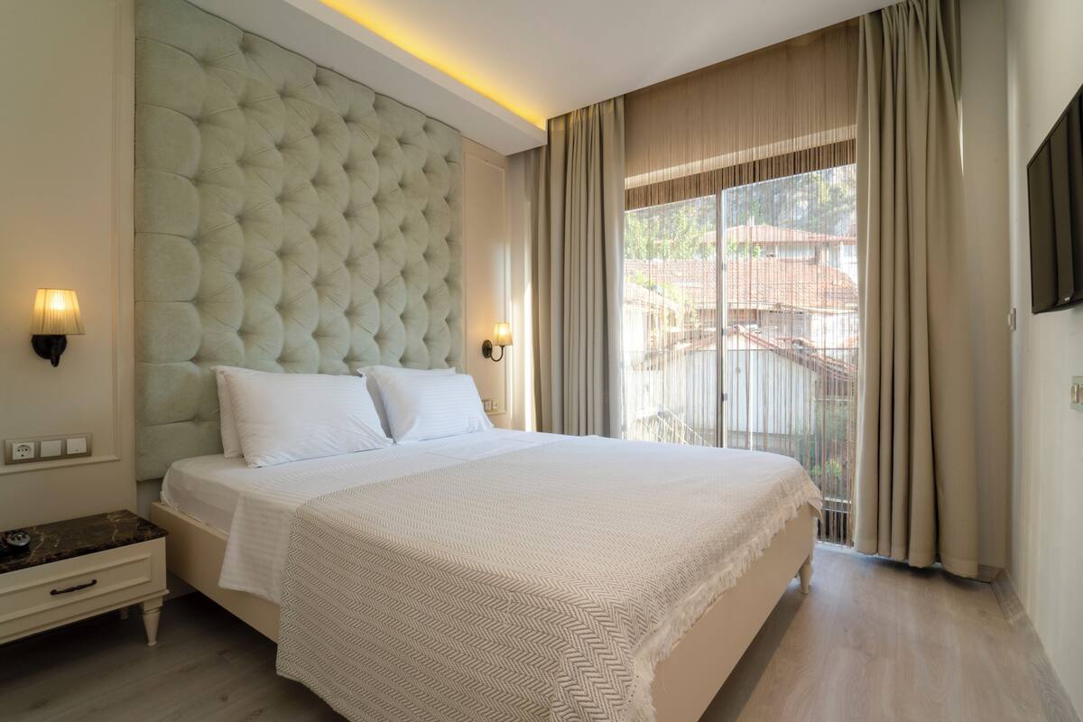 A hotel room with a large bed, a nightstand, a wall-mounted TV, and a window with curtains.