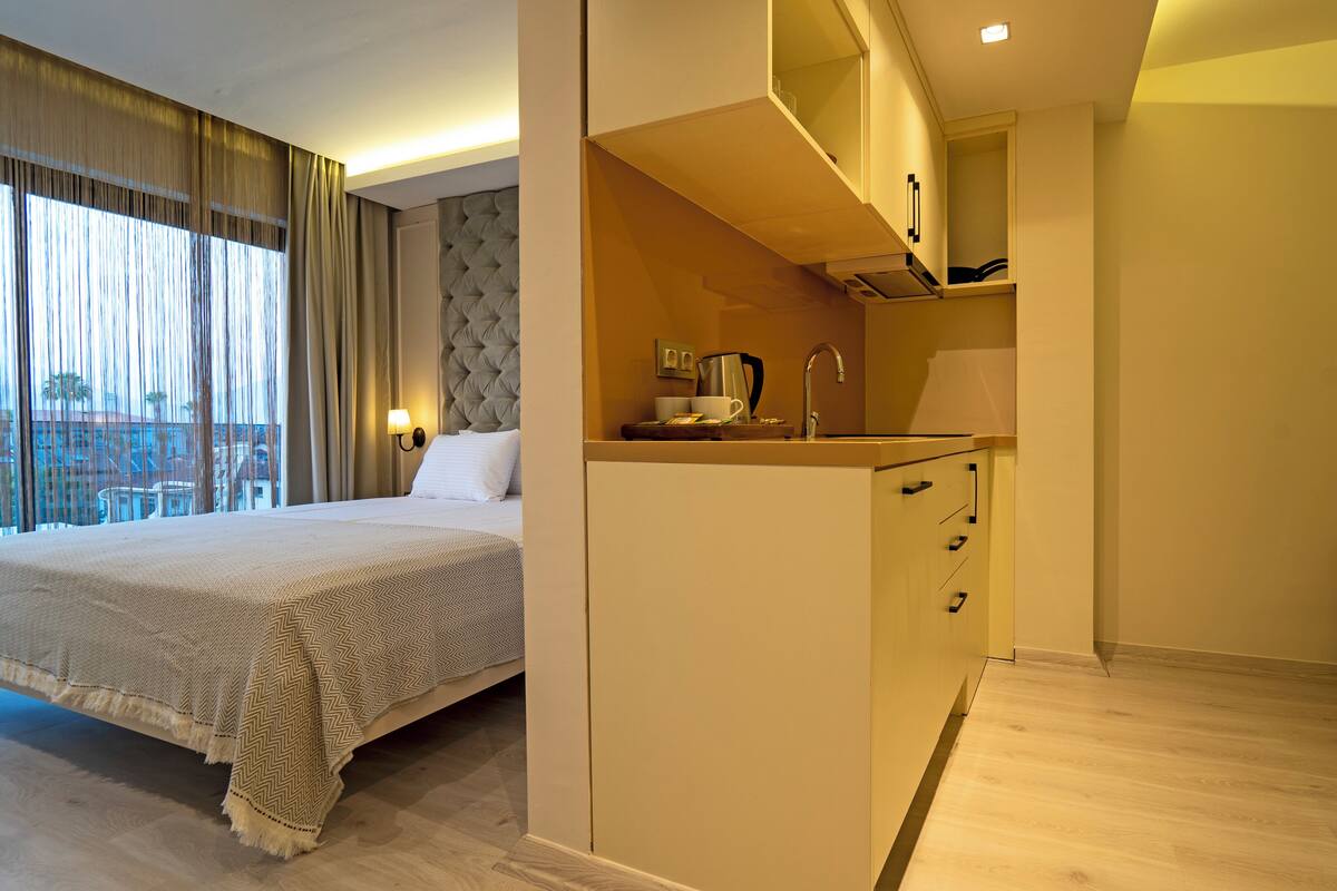 A hotel room with a bed, a kitchenette, a sofa, and a view of buildings outside.