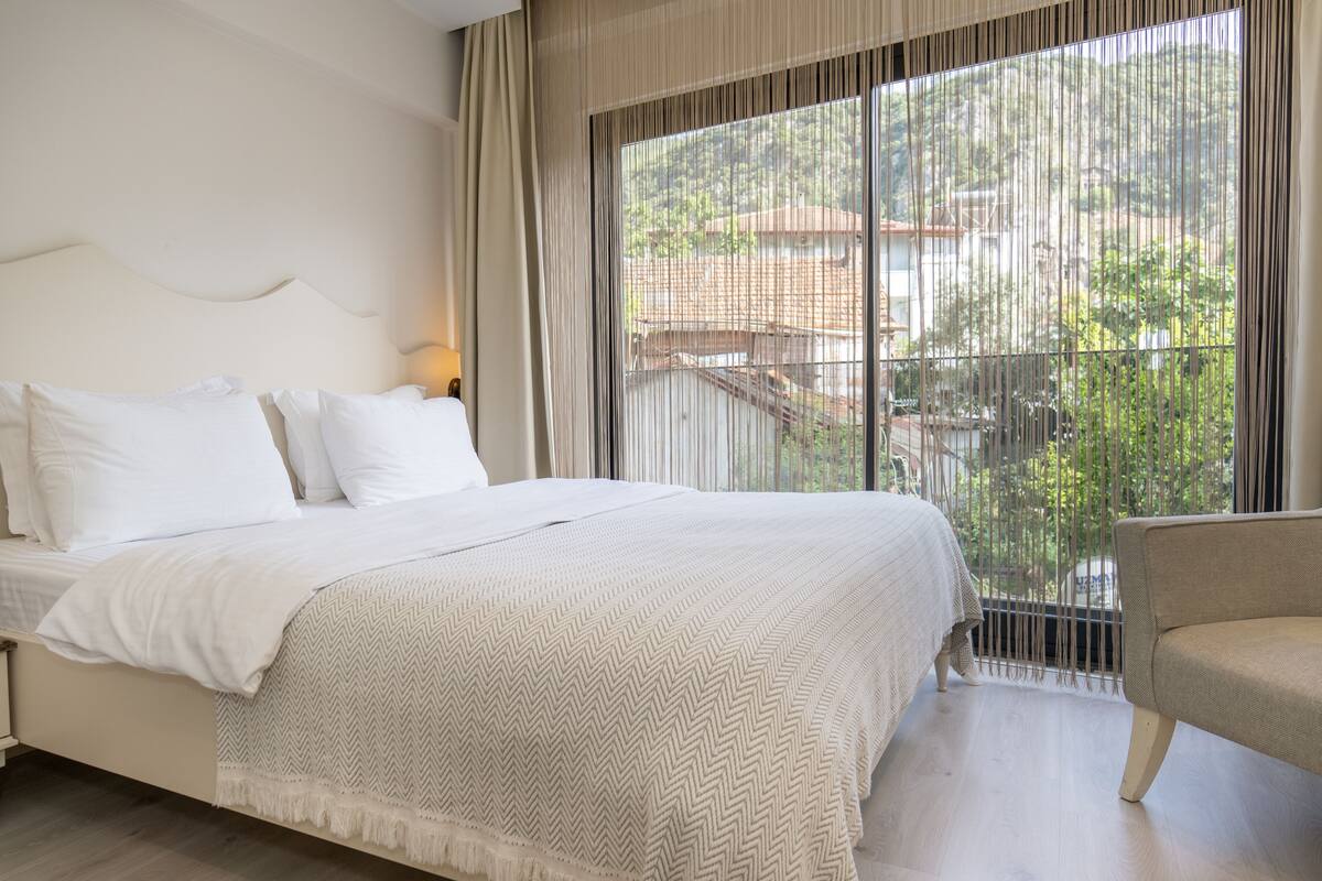 A bedroom with a large bed, a chair, and a view of a balcony with greenery and a building in the background.