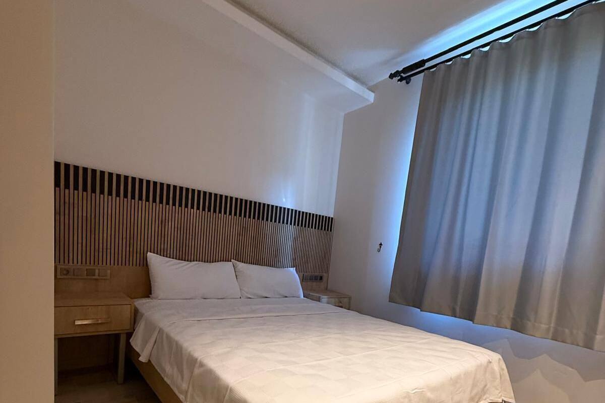 A modern bedroom with a wooden bed, a headboard with vertical slats, a bedside table, and a large window with curtains.