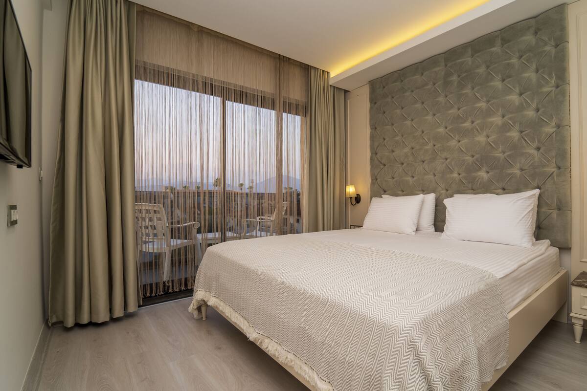 A hotel room with a large bed, a view through the window, and a balcony with chairs.