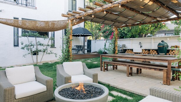 Un patio al aire libre con mobiliario de mimbre, un pozo de fuego y una pérgola con luces colgantes.