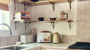 Una cocina con armarios blancos, horno empotrado, cafetera y estantería con libros y objetos decorativos.