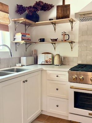 Una cocina con armarios blancos, horno empotrado, cafetera y estantería con libros y objetos decorativos.