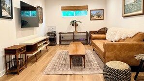 Una sala de estar con piso de madera, un sofá marrón, una mesa de centro, un televisor montado en la pared y obras de arte enmarcadas.