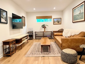 Una sala de estar con piso de madera, un sofá marrón, una mesa de centro, un televisor montado en la pared y obras de arte enmarcadas.