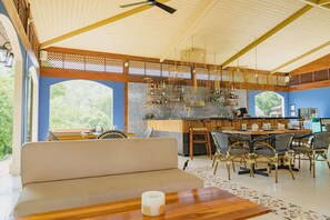A spacious room with a high ceiling, a bar area with shelves of bottles, a seating area with a sofa and dining table, and large windows offering a view of greenery. - Paseo Miraflores 8 at Las Catalinas (Las Catalinas)