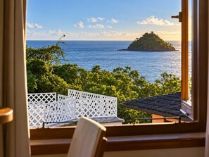 A view from a window overlooking a coastal landscape with a small island, lush greenery, and a clear sky.