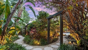 Un sendero en un jardín con piedras circulares, rodeado de exuberante vegetación tropical y una estructura de pérgola de madera.