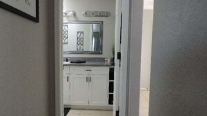 Un pasillo estrecho que conduce a una cocina con armarios blancos, una encimera y un espejo grande sobre el lavabo.