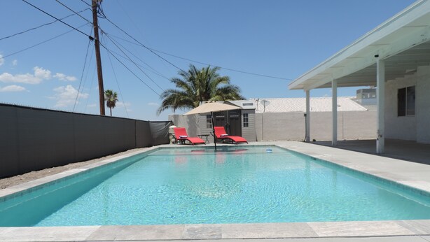 Una piscina rectangular con agua azul cristalina, rodeada por una solana de hormigón. Hay dos sillas reclinables rojas y una sombrilla color arena cerca de la piscina.