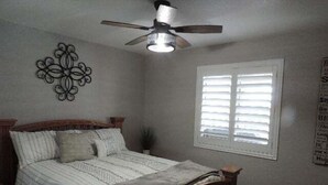 Un dormitorio con una cama, un ventilador de techo, una ventana con persianas y un adorno en la pared.