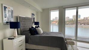 A modern bedroom with a large bed, a nightstand, a dresser, and a view of a cityscape through large windows. - Luxury Vacation Rental Waterfront Condo Bal Harbour Miami Beach (Bay Harbor Islands)