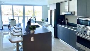 A modern kitchen with a central island, stainless steel appliances, and a view of the city through large windows. - Luxury Vacation Rental Waterfront Condo Bal Harbour Miami Beach (Bay Harbor Islands)