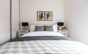 Une chambre à coucher moderne avec un grand lit, deux tables de chevet avec des lampes et deux œuvres d’art abstraites encadrées accrochées au mur.