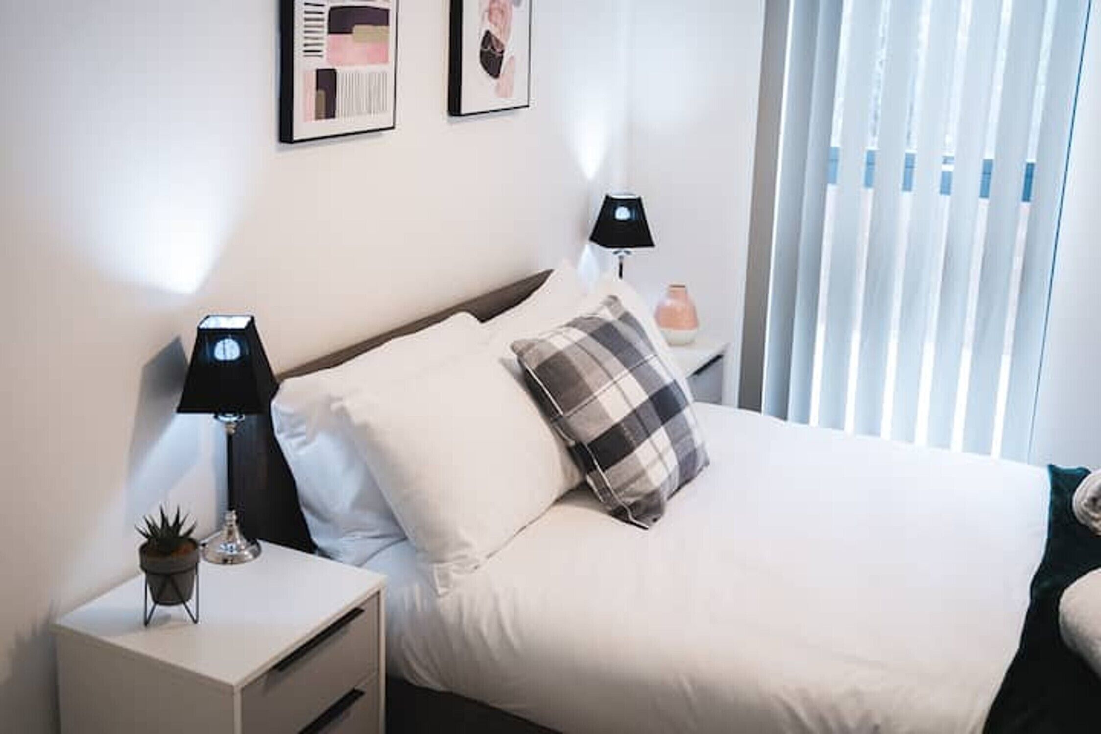 A neatly made bed with white bedding and a plaid pillow, a bedside lamp, a small potted plant, and a framed picture on the wall.