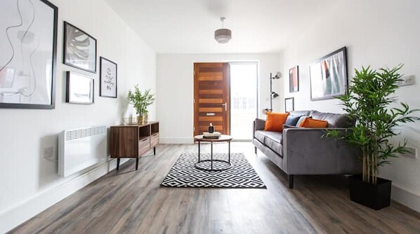 Un salon moderne avec un parquet, un canapé gris agrémenté de coussins orange, un tapis noir et blanc, et une porte en bois à l’arrière-plan.